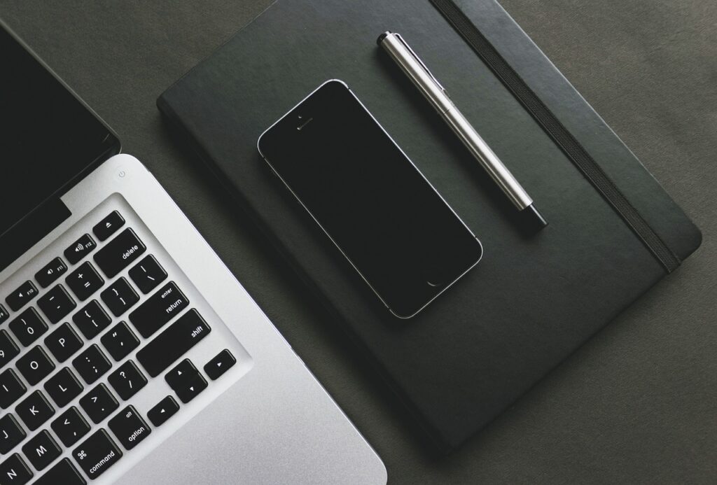 A flat lay of tech essentials including a laptop, a smartphone, a black notebook, and a silver pen arranged on a dark surface.